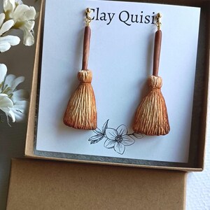 Puede incluir: Un par de pendientes de escoba marrones y dorados en una peque&ntilde;a caja con el texto "Clay Quisit" en la tapa. La caja es marr&oacute;n con una etiqueta blanca que dice "The Clay Quisit" y tiene un gr&aacute;fico de mariposa.