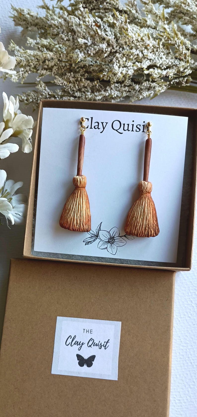 Puede incluir: Un par de pendientes de escoba marrones y dorados en una peque&ntilde;a caja con el texto "Clay Quisit" en la tapa. La caja es marr&oacute;n con una etiqueta blanca que dice "The Clay Quisit" y tiene un gr&aacute;fico de mariposa.