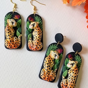 May include: Two pairs of dangle earrings featuring a rectangular black base with a sculpted orange and black spotted leopard sitting among green leaves and colorful tropical flowers. The earrings have gold-toned metal hooks and black circular studs.