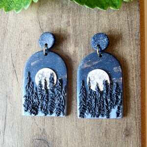 May include: A pair of dark blue and silver polymer clay earrings with a moon and forest scene. The earrings are shaped like a rounded rectangle and have a textured surface.