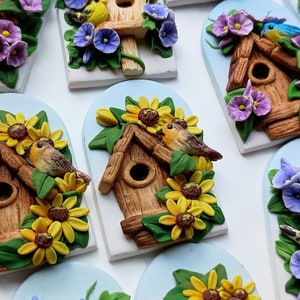May include: A collection of handmade birdhouse ornaments with various floral accents. Each ornament features a small wooden birdhouse with a hole for a bird to enter, surrounded by colorful flowers and leaves. The birdhouses are painted in shades of brown and beige, while the flowers are in shades of purple, yellow, and green.