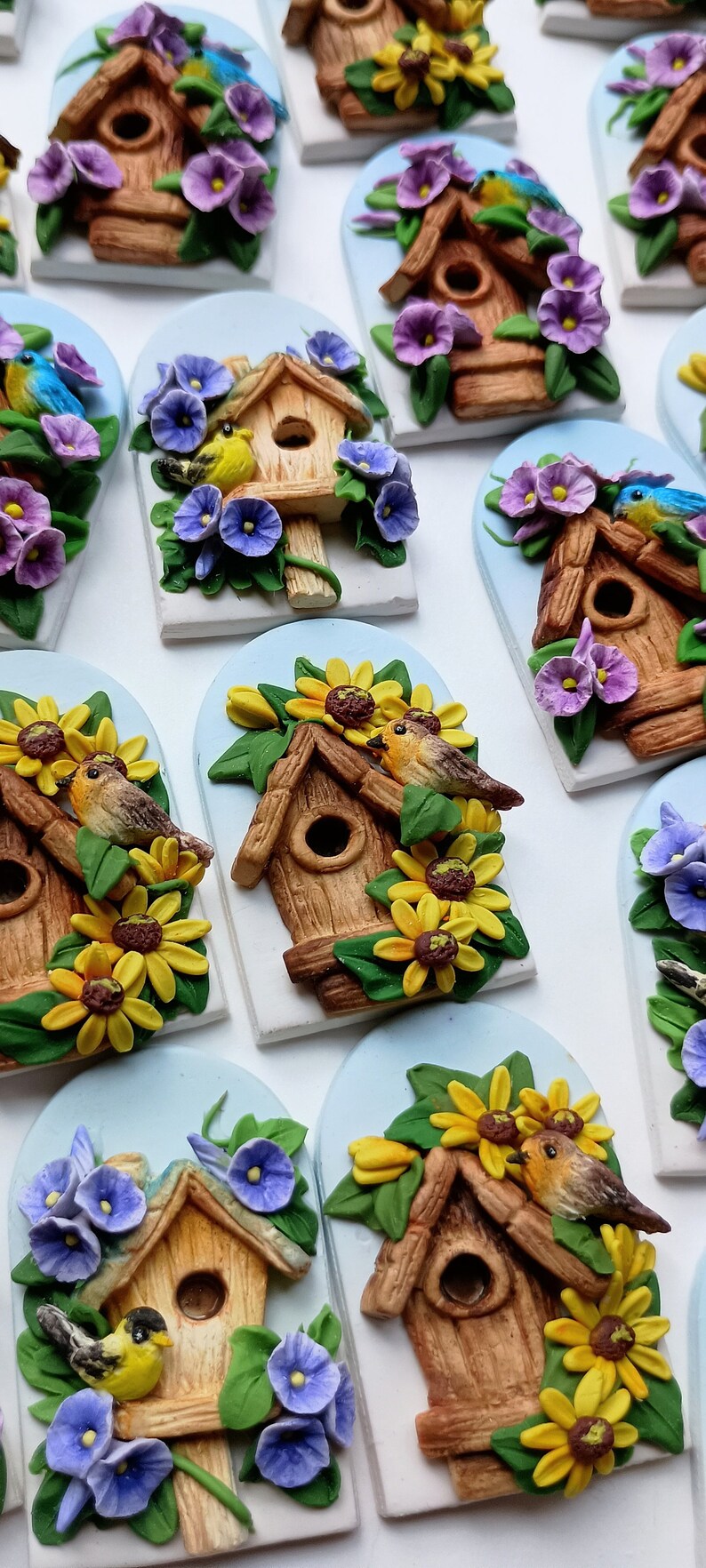 May include: A collection of handmade birdhouse ornaments with various floral accents. Each ornament features a small wooden birdhouse with a hole for a bird to enter, surrounded by colorful flowers and leaves. The birdhouses are painted in shades of brown and beige, while the flowers are in shades of purple, yellow, and green.