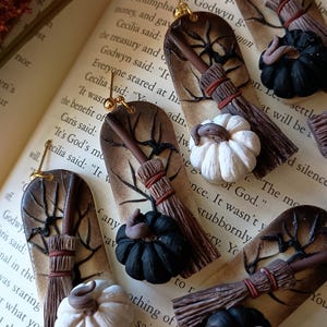 May include: A pair of handmade Halloween-themed earrings featuring a brown and white broom, a black and white pumpkin, and a brown tree branch with a spider web. The earrings are made of polymer clay and have gold hooks.