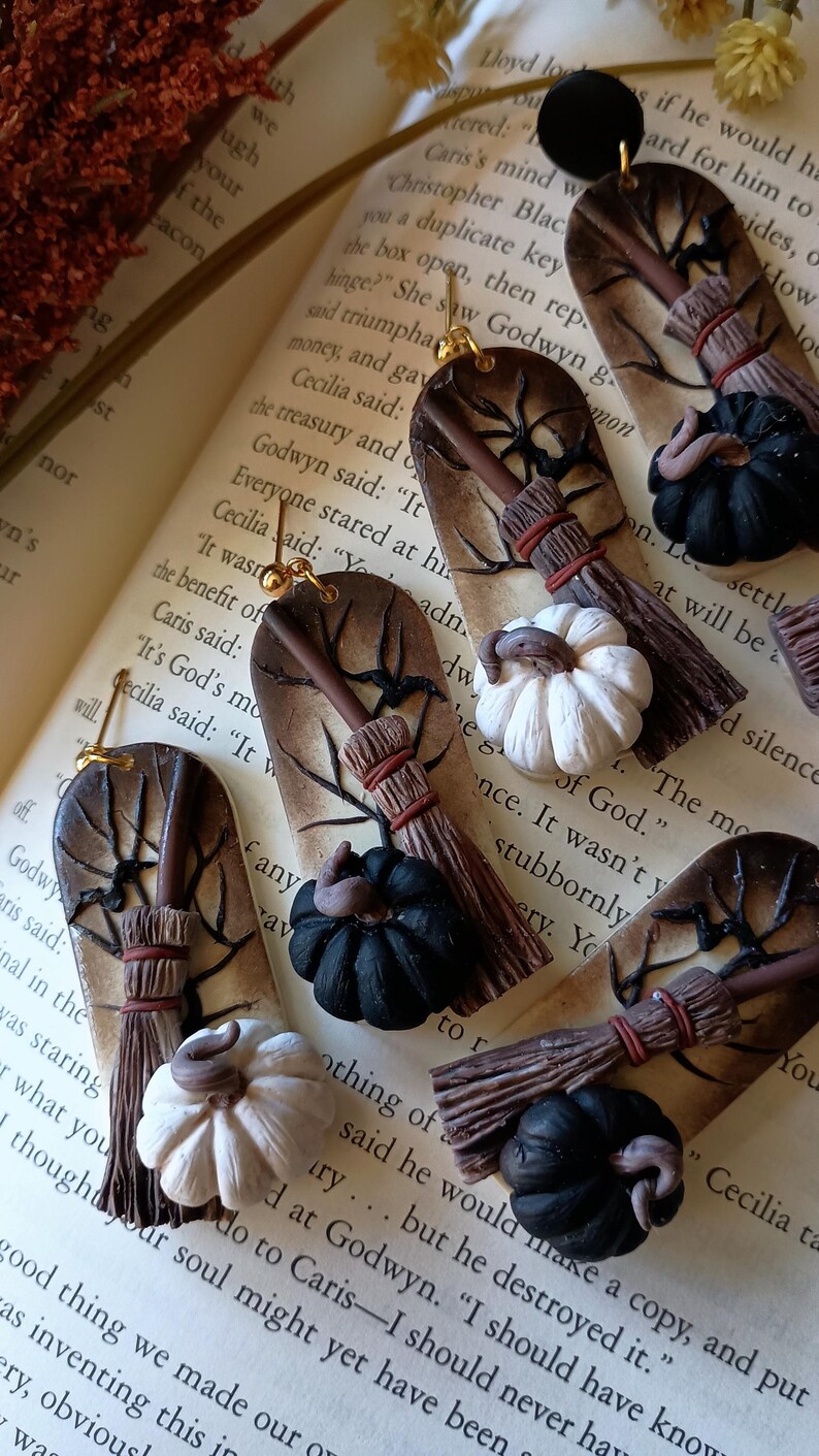 May include: A pair of handmade Halloween-themed earrings featuring a brown and white broom, a black and white pumpkin, and a brown tree branch with a spider web. The earrings are made of polymer clay and have gold hooks.