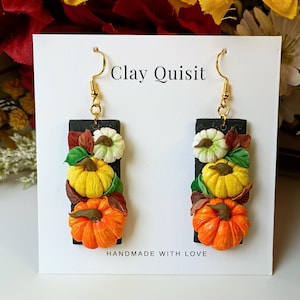 May include: Rectangular black earrings featuring sculpted white, yellow, and orange pumpkins with green leaves and brown stems. The earrings hang from gold-toned hooks on a white display card labeled Clay Quisit and Handmade with Love.