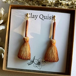 Puede incluir: Un par de pendientes hechos a mano de arcilla con forma de escobas. Los pendientes son de color marr&oacute;n claro y tienen un gancho dorado. Los pendientes se muestran en una tarjeta blanca con el texto "Clay Quisi" impreso en ella.