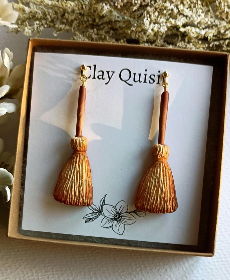 Puede incluir: Un par de pendientes hechos a mano de arcilla con forma de escobas. Los pendientes son de color marr&oacute;n claro y tienen un gancho dorado. Los pendientes se muestran en una tarjeta blanca con el texto "Clay Quisi" impreso en ella.