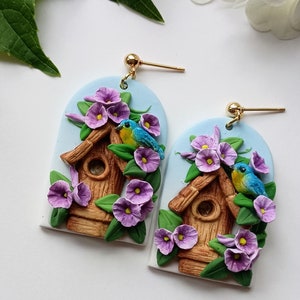 May include: A pair of earrings featuring a birdhouse with two blue birds perched on the edge, surrounded by purple flowers. The earrings are made of polymer clay and have gold hooks.