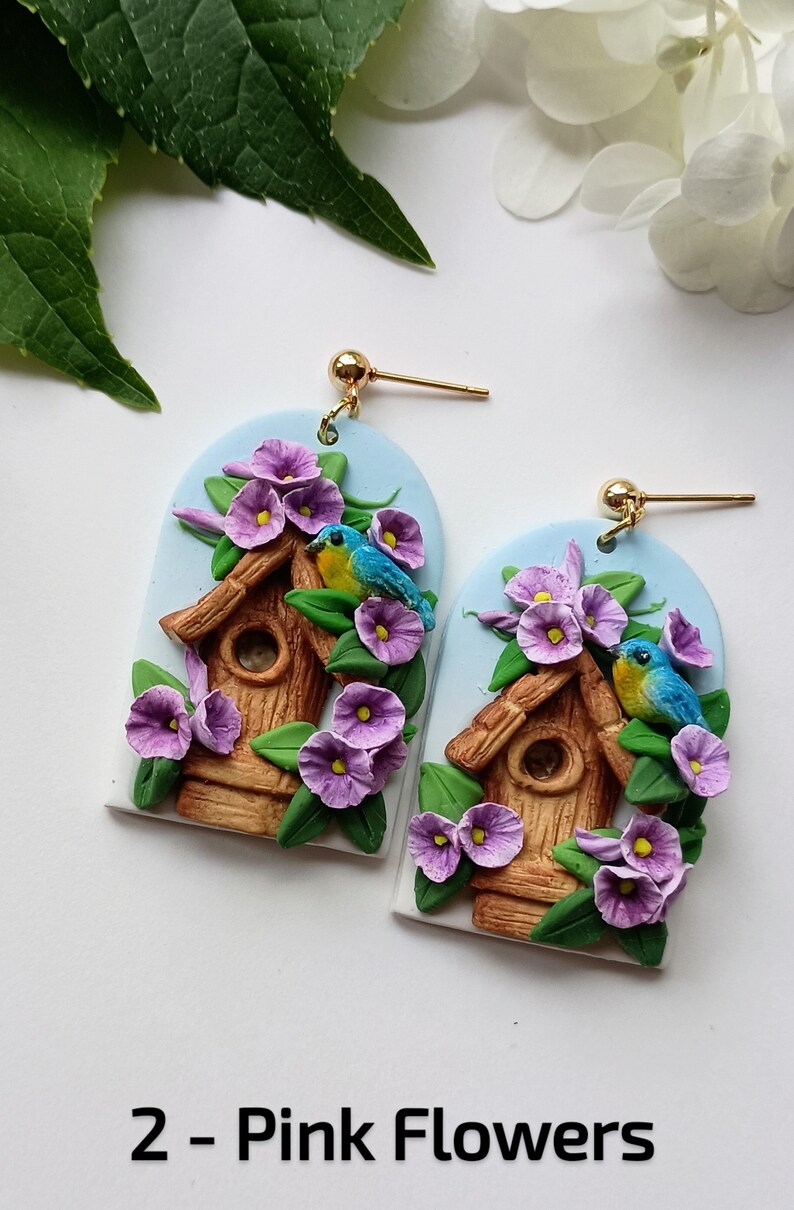 May include: A pair of earrings featuring a birdhouse with two blue birds perched on the edge, surrounded by purple flowers. The earrings are made of polymer clay and have gold hooks.