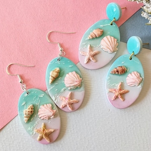 May include: Oval-shaped earrings with a beach theme. Each earring features a light blue and pink background with seashells and starfish. The earrings have silver hooks and a turquoise stud at the top.