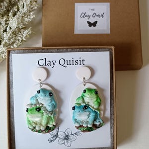 May include: A pair of frog-themed earrings with a white background. The earrings are made of polymer clay and feature three frogs stacked on top of each other. The frogs are painted in shades of green and blue. The earrings are packaged in a white box with the text "Clay Quisit" printed on the front.