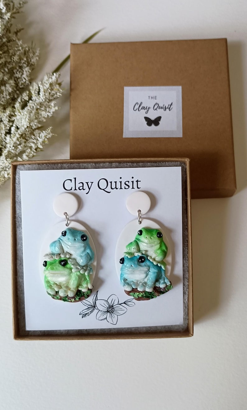 May include: A pair of frog-themed earrings with a white background. The earrings are made of polymer clay and feature three frogs stacked on top of each other. The frogs are painted in shades of green and blue. The earrings are packaged in a white box with the text "Clay Quisit" printed on the front.