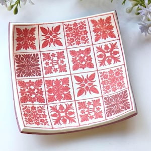 Op de afbeelding: Vierkante keramische schaal met een witte basis en een rood bloemmotief. De schaal is verdeeld in zestien vierkanten, elk met een ander bloemdesign. De randen van de schaal zijn donkerrood.