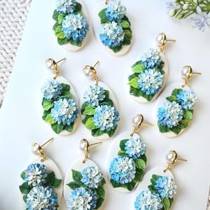 May include: Oval earrings with blue and white hydrangea flower clusters and green leaves. Each earring has a gold-coloured post and a pearl accent. The earrings are set against a white background.