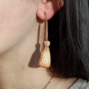 Puede incluir: Un par de pendientes de color dorado con un peque&ntilde;o amuleto de escoba. La escoba est&aacute; hecha de material marr&oacute;n y dorado y tiene un mango largo y delgado.