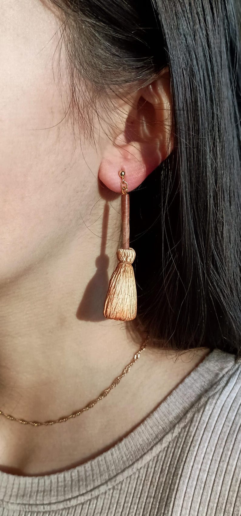 Puede incluir: Un par de pendientes de color dorado con un peque&ntilde;o amuleto de escoba. La escoba est&aacute; hecha de material marr&oacute;n y dorado y tiene un mango largo y delgado.