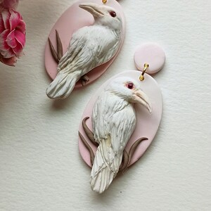 May include: A pair of white bird earrings with red eyes, perched on pink oval-shaped bases. The earrings are made of polymer clay and have a delicate, whimsical design.