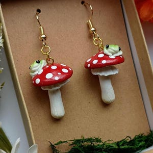 May include: A pair of whimsical earrings featuring red mushroom caps with white polka dots, each topped with a small green frog. The earrings have gold-colored hooks and are displayed in a brown box.