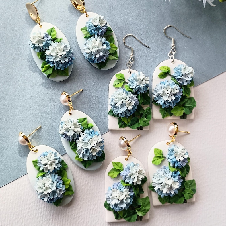 May include: A selection of handmade earrings with intricate floral designs. The earrings display white, blue, and green hydrangea flowers. Some are oval, others arched. Gold-tone hooks and pearl details are included. The earrings are suitable for various occasions.