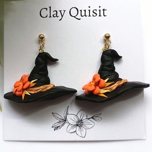 May include: A pair of black witch hat earrings with orange flowers and gold-colored accents. The earrings are handmade and feature a whimsical design, perfect for Halloween or a unique accessory. The earrings are displayed on a white card with the text "Clay Quisit".