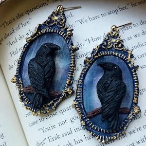 May include: A pair of oval-shaped earrings featuring a three-dimensional black raven perched on a brown branch. The raven is set against a blue and purple gradient background, enclosed in a dark blue frame with gold-colored highlights and ornate detailing.