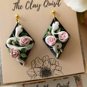 May include: A pair of black diamond-shaped earrings featuring white snakes and pink roses. The earrings are handmade polymer clay jewellery by The Clay Quisit.