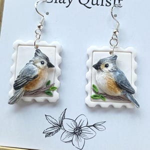 Op de afbeelding: Handgemaakte oorbellen met vogelsculpturen. Elke oorbel toont een vogel met grijs en bruin verenkleed, zittend op een tak met groene bladeren, geplaatst in een witte, postzegelvormige lijst. De oorbellen hangen aan zilverkleurige haken.