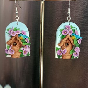 May include: A pair of bluebird earrings with a birdhouse and purple flowers. The earrings are made of polymer clay and have silver hooks.