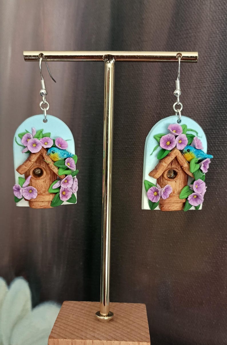 May include: A pair of bluebird earrings with a birdhouse and purple flowers. The earrings are made of polymer clay and have silver hooks.