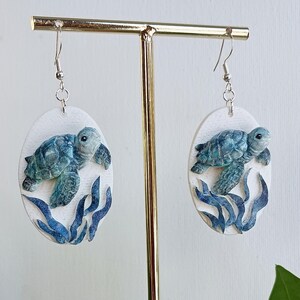 Puede incluir: Un par de pendientes colgantes ovalados con un fondo blanco texturizado y una tortuga marina tridimensional en relieve de tonos azules y verdes nadando sobre algas de color azul oscuro. Los pendientes cuelgan de ganchos de metal plateado.