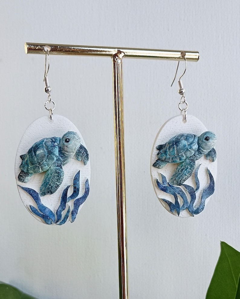 Puede incluir: Un par de pendientes colgantes ovalados con un fondo blanco texturizado y una tortuga marina tridimensional en relieve de tonos azules y verdes nadando sobre algas de color azul oscuro. Los pendientes cuelgan de ganchos de metal plateado.