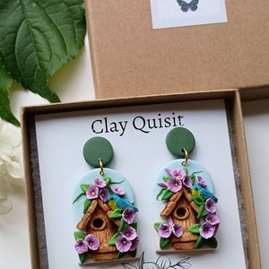 May include: A pair of polymer clay earrings featuring a birdhouse with blue birds and purple flowers. The earrings are oval shaped and have a green clay backing. The earrings are packaged in a box with the text "Clay Quisit" on the front.