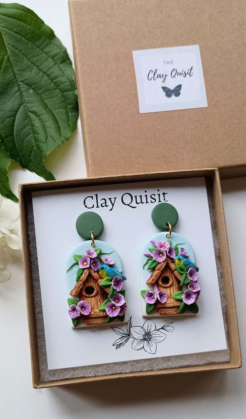 May include: A pair of polymer clay earrings featuring a birdhouse with blue birds and purple flowers. The earrings are oval shaped and have a green clay backing. The earrings are packaged in a box with the text "Clay Quisit" on the front.
