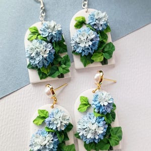 May include: Four pairs of earrings with a floral design. Each earring features a white rectangular base with a blue and white hydrangea flower and green leaves. Two pairs have silver hooks, and two have gold posts with pearls.