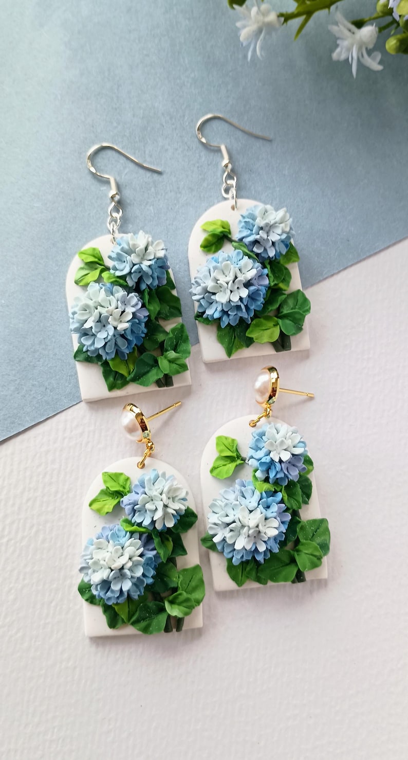 May include: Four pairs of earrings with a floral design. Each earring features a white rectangular base with a blue and white hydrangea flower and green leaves. Two pairs have silver hooks, and two have gold posts with pearls.