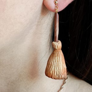 Puede incluir: Un par de pendientes de oro con un amuleto de escoba marr&oacute;n y dorado. La escoba est&aacute; hecha de un material texturizado que se asemeja a la paja.