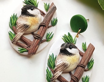 May include: A pair of handcrafted earrings featuring a detailed bird design. Each earring showcases a small bird perched on a brown branch, surrounded by green foliage, set against a white oval backdrop. The earrings have green stud tops.