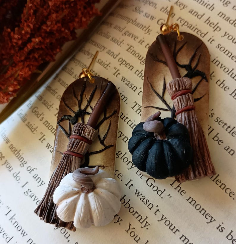 May include: A pair of handmade polymer clay earrings featuring a witch's broom, a black pumpkin, and a white pumpkin. The earrings are attached to gold ear wires.