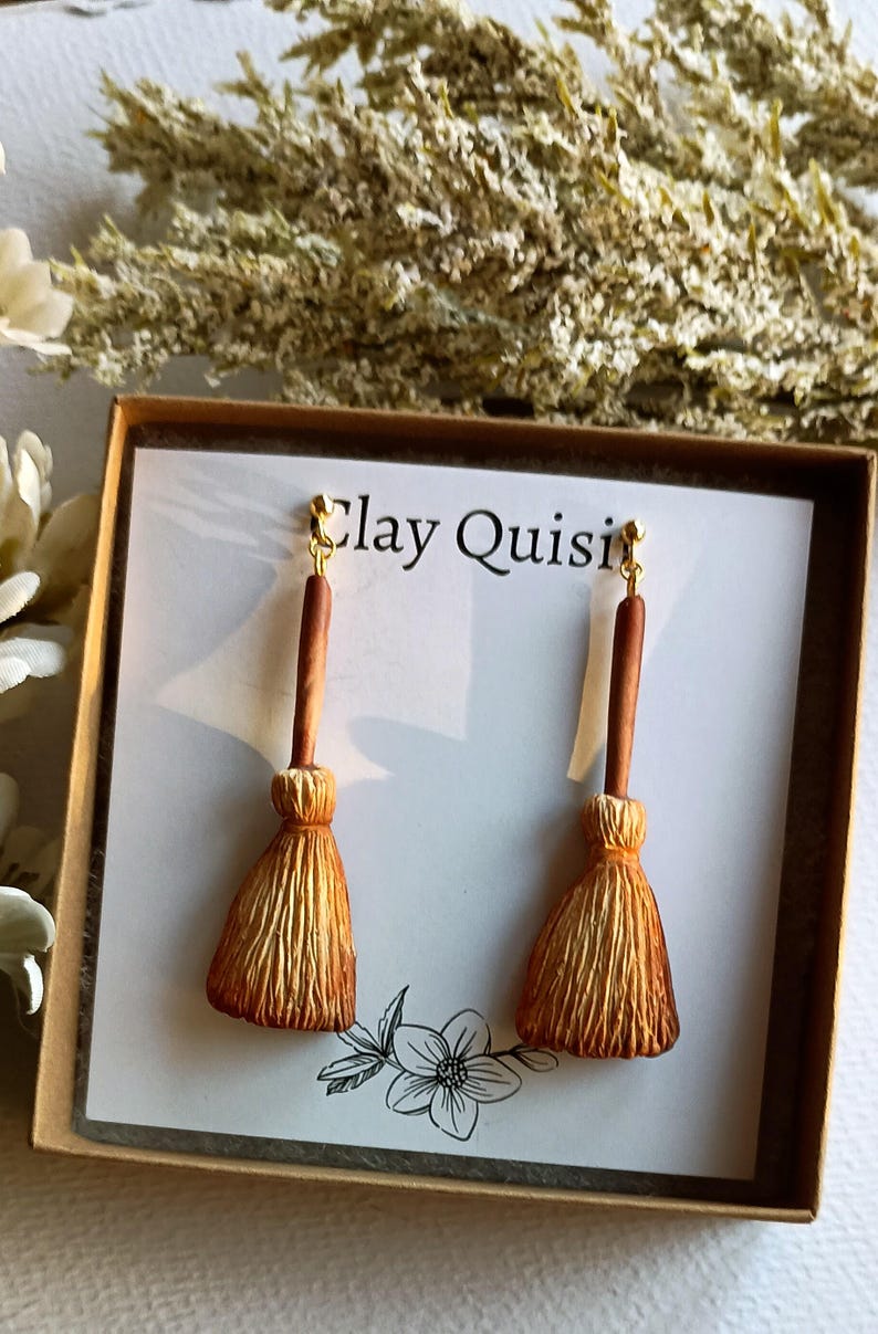 Puede incluir: Un par de pendientes de escoba marrones y dorados en una caja blanca con el texto "Clay Quisi" en la parte delantera. Los pendientes est&aacute;n hechos de arcilla polim&eacute;rica y tienen un aspecto realista.