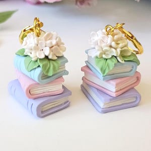May include: A pair of earrings featuring stacks of three miniature books in pastel blue, pink, and purple. Each stack is topped with a cluster of white hydrangea flowers and green leaves, finished with a gold-toned metal earring hook attachment.