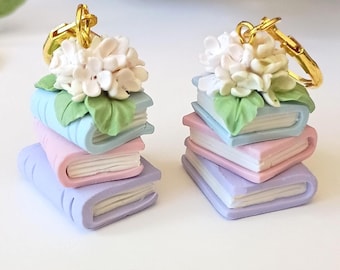 Könnte beinhalten: Ein Paar Ohrringe mit Stapeln aus drei Miniaturbüchern in Pastellblau, Rosa und Lila. Jeder Stapel ist mit einem Büschel weißer Hortensienblüten und grünen Blättern verziert und mit einem goldfarbenen Metall-Ohrhaken versehen.