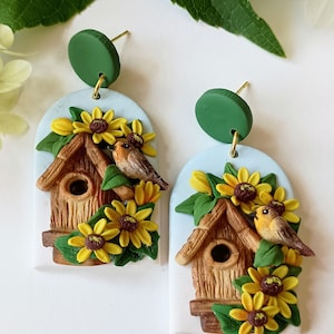 Puede incluir: Un par de pendientes hechos a mano de arcilla polimérica con una casita de pájaros con techo marrón y un agujero redondo, rodeada de girasoles amarillos y hojas verdes. Cada pendiente tiene un pequeño círculo verde en la parte superior y un gancho dorado.