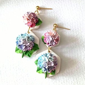 Könnte beinhalten: Handgefertigte Ohrringe mit Miniatur-Hortensienblüten in Rosa, Lila und Blau. Die floralen Cluster sind an weißen, sechseckigen Anhängern mit goldfarbenen Ohrhaken befestigt. Grüne Blätter akzentuieren die Blumen.