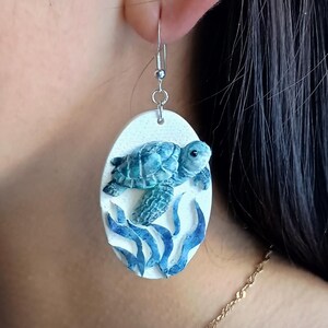 Puede incluir: Un pendiente colgante de forma ovalada con un fondo blanco texturizado, que presenta una tortuga marina de color verde azulado esculpida nadando sobre algas azules. El pendiente cuelga de un gancho de metal plateado y muestra un dise&ntilde;o en relieve.