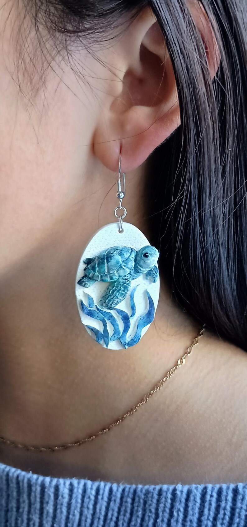 Puede incluir: Un pendiente colgante de forma ovalada con un fondo blanco texturizado, que presenta una tortuga marina de color verde azulado esculpida nadando sobre algas azules. El pendiente cuelga de un gancho de metal plateado y muestra un dise&ntilde;o en relieve.