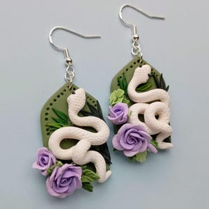 May include: Handmade earrings with a white snake design on a green background. The earrings are decorated with purple roses and green leaves, with silver-coloured ear wires.