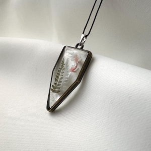 Può includere: Una collana con un ciondolo d'argento e un ciondolo in resina trasparente con una felce pressata e un piccolo fiore rosa. Il ciondolo è incastonato in una cornice geometrica di colore bronzo.