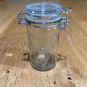 May include: A clear glass jar with a hinged lid and a metal clasp. The jar features a decorative etched design of a stylized shark's jaw. The jar is empty and sits on a wooden surface.