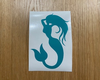 May include: A teal mermaid silhouette sticker. The mermaid has flowing hair and a fishtail. The sticker is on a white background, with a wooden surface in the background.