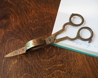 May include: Antique brass scissors with an ornate design and a pointed tip. The scissors are closed and lying on a notebook with blue ink writing.